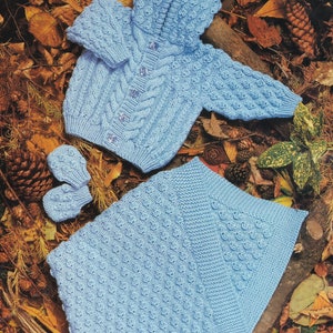 May include: A light blue knitted baby set with a hooded cardigan, matching mittens, and a blanket. The cardigan has a cable knit pattern and buttons down the front. The blanket and mittens have a textured stitch.