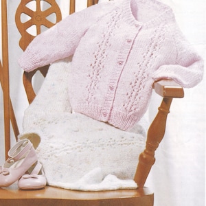 May include: A pink knitted cardigan with a button-up front and a delicate lace pattern. The cardigan is draped over a wooden chair with a white knitted blanket.