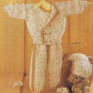 May include: A cream-colored knitted baby outfit, including a jacket, pants, and hat, with brown trim. The jacket has buttons and a shawl collar. The outfit is displayed with a teddy bear and a wicker basket.