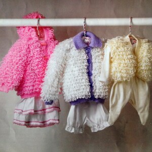 May include: Three crocheted baby sweaters hanging on a white rack. The sweaters are pink, white, and yellow. The pink sweater has a white ruffle skirt attached. The white sweater has a purple button-up shirt underneath. The yellow sweater has a white pair of pants attached.