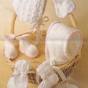 May include: A set of white knitted baby clothes, including a bonnet, booties, and mittens. The items are arranged in a wicker basket with a white ribbon.