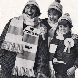 Op de afbeelding: Een gezin van drie personen draagt witte en zwarte gestreepte sjaals met de tekst "Up for the Cup". Het gezin draagt wintermutsen en glimlacht.