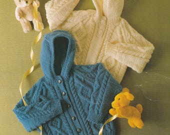 May include: Two knitted hooded cardigans, one teal and one cream, with cable knit patterns. A small teddy bear and a yellow dog toy are placed near the garments. The cardigans have button fastenings.
