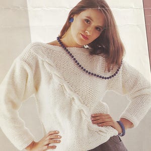 以下が含まれることがあります： ボートネックとケーブル編みのデザインが施された白いニットセーター。長い茶色の髪と青いイヤリングをした人が着用しています。その人は濃い青色のビーズのネックレスをしています。