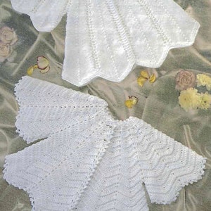 Könnte beinhalten: Zwei weiße gehäkelte Baby-Kleidungsstücke, eines mit einem gewellten Rand und das andere mit einem welligen Rand. Beide Kleidungsstücke liegen auf einem strukturierten Stoffhintergrund.