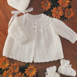 May include: A white knitted baby outfit set including a cardigan, hat, and booties. The set is laid out on a brown wooden surface with orange flowers.