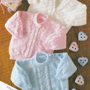 May include: Three knitted baby sweaters in pink, white, and light blue. The sweaters have a delicate cable stitch pattern and button closures. The sweaters are laid out on a wooden surface with decorative hearts.