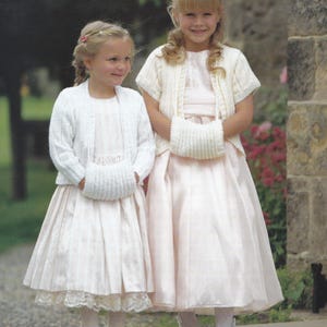 May include: Two young girls wearing white dresses and matching white knitted cardigans with matching white knitted mittens. The girls are standing in front of a stone wall.