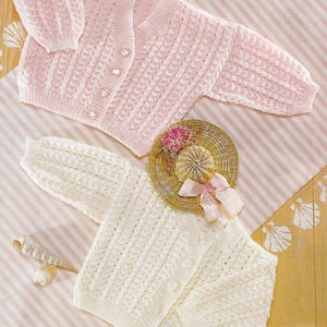 以下が含まれることがあります： 繊細な透かし編みのパターンが施された、ピンクと白の2つのベビー用ニットカーディガン。カーディガンは、貝殻が置かれたストライプの背景の上に置かれています。