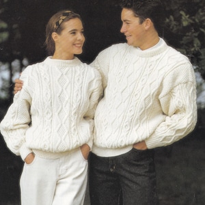 Könnte beinhalten: Zwei Personen tragen weiße Strickpullover mit Zopfmuster. Die Person links trägt eine weiße Hose und die Person rechts trägt eine dunkelblaue Jeans.