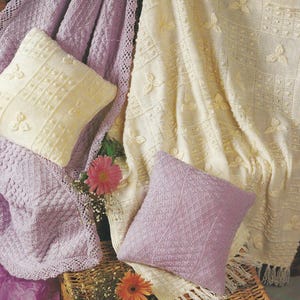 May include: A cozy scene featuring knitted blankets and pillows in soft pastel colors. The blankets are cream and lavender, with intricate patterns and textures. Two pillows are cream and lavender, adding to the comfort. The scene is set on a wicker basket.