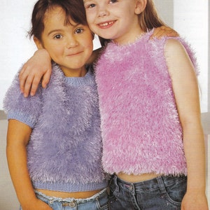 Puede incluir: Dos chicas jóvenes con tops de punto peludo y vaqueros. Una lleva un jersey lila de manga corta y la otra un top rosa sin mangas. Sonríen y se abrazan. Los tops tienen un aspecto suave y texturizado.