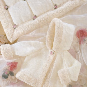 May include: A white knitted baby jacket with a hood and a button closure. The jacket has a fluffy white trim around the edges and a small pink rose button.