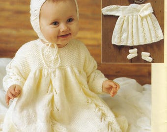 May include: A baby wearing a cream-colored knitted outfit, including a bonnet, dress, and booties. The dress features a textured pattern and long sleeves. A matching set of baby clothes is displayed on a wooden surface.