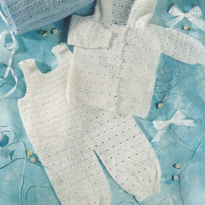 Op de afbeelding: Een witte gehaakte baby-outfitset met een capuchonjack, broek en een bijpassend topje zonder mouwen. De set ligt op een blauwe achtergrond met witte linten en kleine witte bloemen.