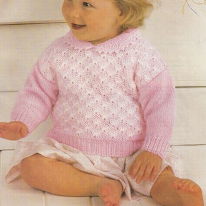 May include: A pink knitted baby sweater with a white lace pattern. The sweater has a collar and long sleeves.