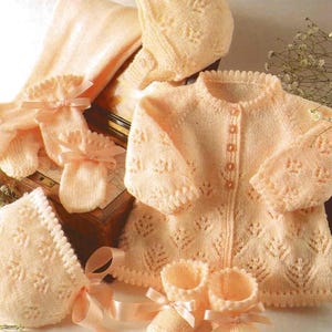Puede incluir: Un conjunto de ropa de bebé tejido en color melocotón pálido. El conjunto incluye un cárdigan, un gorro, botines y manoplas. El cárdigan tiene un delicado patrón de encaje y se abrocha con botones en la parte delantera. El gorro y los botines también están decorados con encaje.