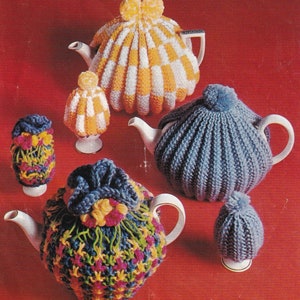 Puede incluir: Cinco cubiertas de tetera de punto en varios colores y patrones. Las cubiertas están hechas de hilo y presentan diferentes texturas y diseños. Las cubiertas se muestran sobre un fondo rojo.