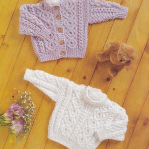 May include: Two knitted baby sweaters, one in white and one in lavender, with a cable knit pattern. The sweaters are laid out on a wooden surface.