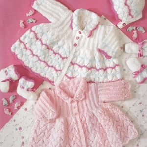 Könnte beinhalten: Zwei gestrickte Babyjacken, eine weiße mit rosa und blauen Akzenten und eine rosa mit weißem Kragen. Beide Jacken haben einen Knopfverschluss und sind mit einem zarten Stichmuster gearbeitet. Zu den Jacken gehören passende gestrickte Mützen und Booties.