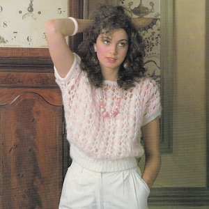 May include: A light pink open-weave knit top with short sleeves. The top is paired with white high-waisted pants. A pink and white beaded necklace and a white bracelet complete the look. The woman is standing in front of a wooden cabinet and a floral wallpaper.