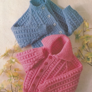 May include: Two knitted cardigans, one blue and one pink, with a cable knit pattern. The cardigans are laid out on a white surface with a branch of leaves.