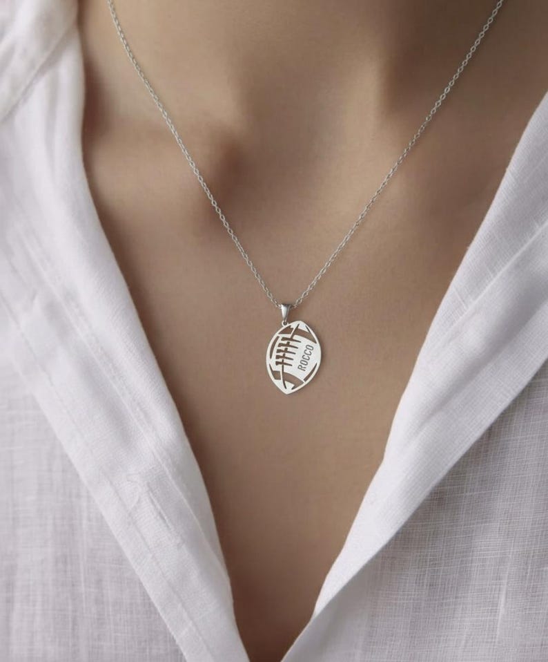 May include: A silver necklace with a football pendant. The pendant is cut out in the shape of a football with the word 'JACK' inscribed inside.