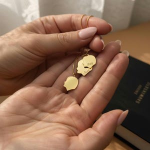 May include: A gold-colored necklace with three pendants. Two pendants are heart-shaped with the names "Ray" and "Alex" engraved on them. The third pendant is a profile of a person's head. The necklace is held in a person's hand.
