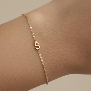 May include: A delicate gold bracelet with the letter "S". The fine chain bracelet features a small, stylised letter charm. The bracelet is worn on a wrist, highlighting its simple elegance.