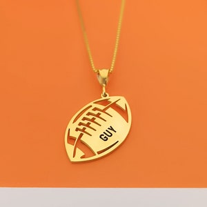 May include: Gold-toned necklace with a football pendant. The pendant is cut out in the shape of a football with the word "GUY" inscribed inside.