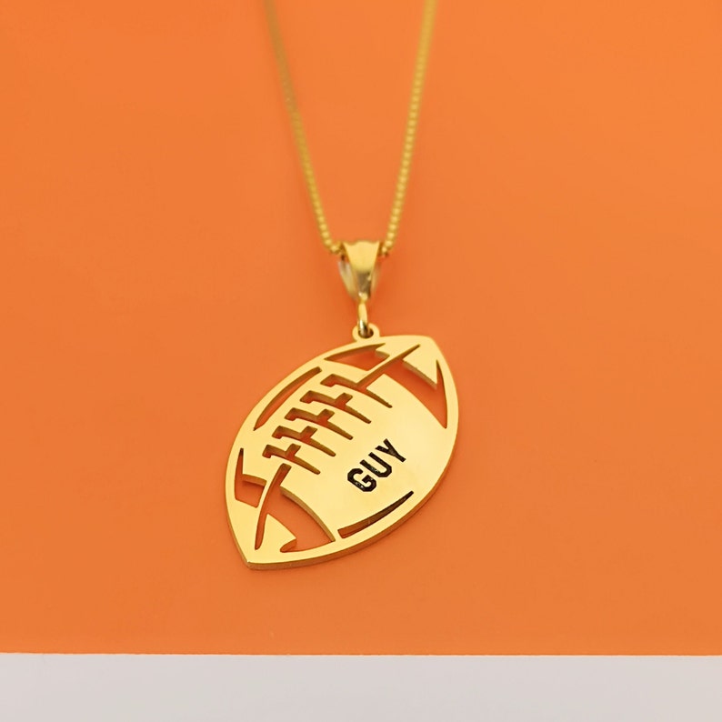May include: Gold-toned necklace with a football pendant. The pendant is cut out in the shape of a football with the word "GUY" inscribed inside.