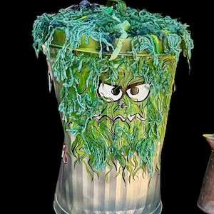 Op de afbeelding: Een groene metalen prullenbak met een groene garen deksel en een getekend cartoon gezicht op de voorkant. Het gezicht heeft grote ogen en een fronsende mond.