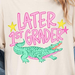 May include: Cream-colored t-shirt with the text "LATER 1ST GRADER" in pink, with yellow stars and a green alligator graphic. The alligator has a pink mouth and is smiling.