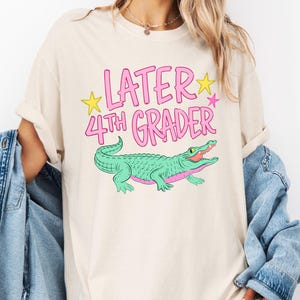 以下が含まれることがあります： クリーム色のTシャツ。ピンク色の「LATER 4TH GRADER」の文字と黄色の星が描かれています。下にはピンクのアクセントが付いた緑色のワニが描かれています。Tシャツの袖は折り返されています。