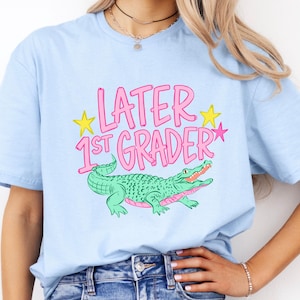Pu&ograve; includere: T-shirt azzurra con la scritta rosa "LATER 1ST GRADER" e stelle gialle. Un alligatore verde con la bocca rosa &egrave; raffigurato sotto. La maglietta &egrave; in stile casual.