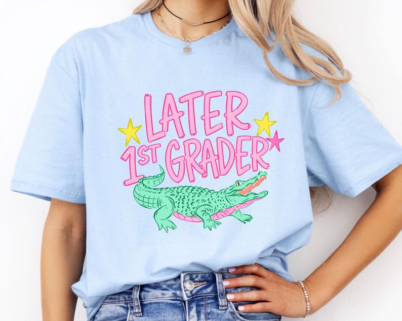 Pu&ograve; includere: T-shirt azzurra con la scritta rosa "LATER 1ST GRADER" e stelle gialle. Un alligatore verde con la bocca rosa &egrave; raffigurato sotto. La maglietta &egrave; in stile casual.
