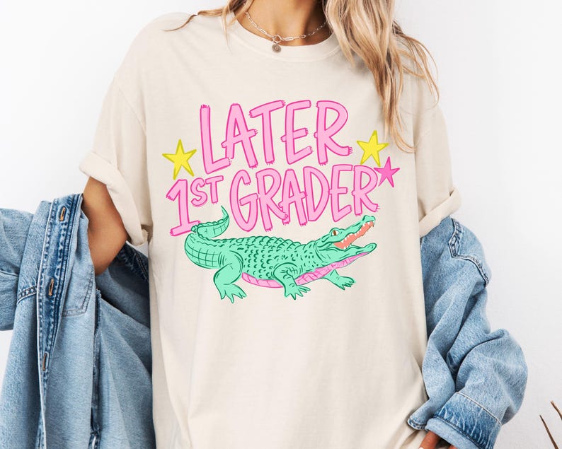 May include: Cream-colored t-shirt with the text "LATER 1ST GRADER" in pink, with yellow stars and a green alligator graphic. The alligator has a pink mouth and is smiling.