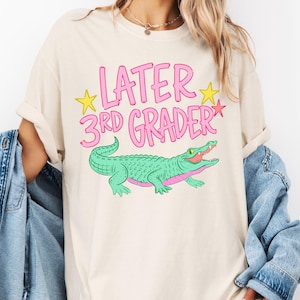 Könnte beinhalten: Cremefarbenes T-Shirt mit dem pinkfarbenen Text "LATER 3RD GRADER", akzentuiert mit gelben Sternen. Ein grünes Alligator-Motiv mit rosa Akzenten befindet sich darunter. Das T-Shirt hat einen lässigen Stil.