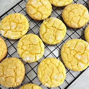 Op de afbeelding: Een bovenaanzicht van gele koekjes op een afkoelrek. De ronde koekjes hebben een gebarsten, getextureerd oppervlak en zijn bestrooid met poedersuiker. Ze zien er vers gebakken uit.