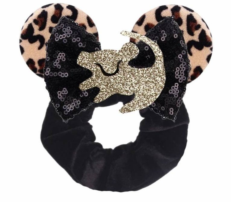 May include: Black velvet hair scrunchie with a black and gold sequined bow and leopard print fabric ears. The bow features a gold glitter lion silhouette.