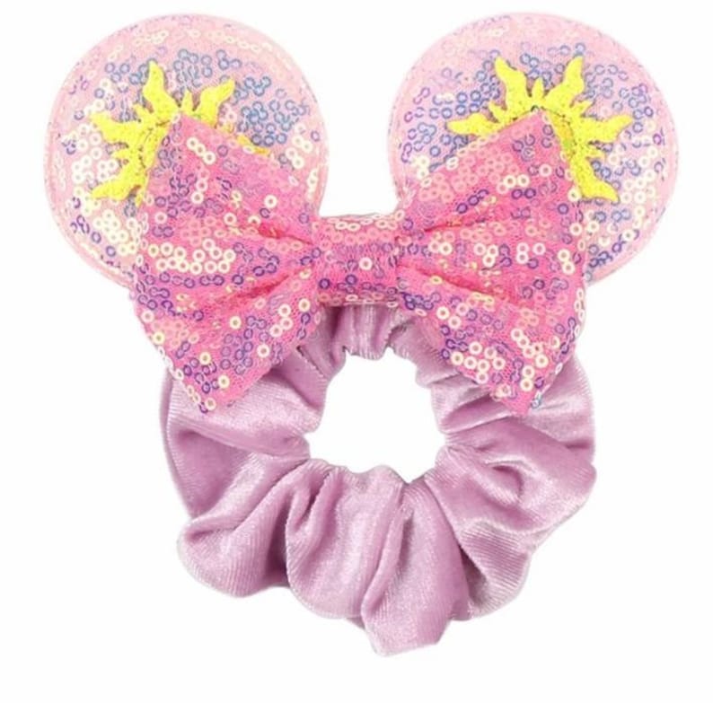 May include: A pink and purple sequined bow with a yellow sun design on each side, attached to a purple velvet scrunchie.