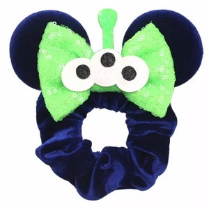 May include: A blue velvet hair scrunchie with a green and white alien-themed bow. The bow has three white eyes with black pupils and a green antenna.