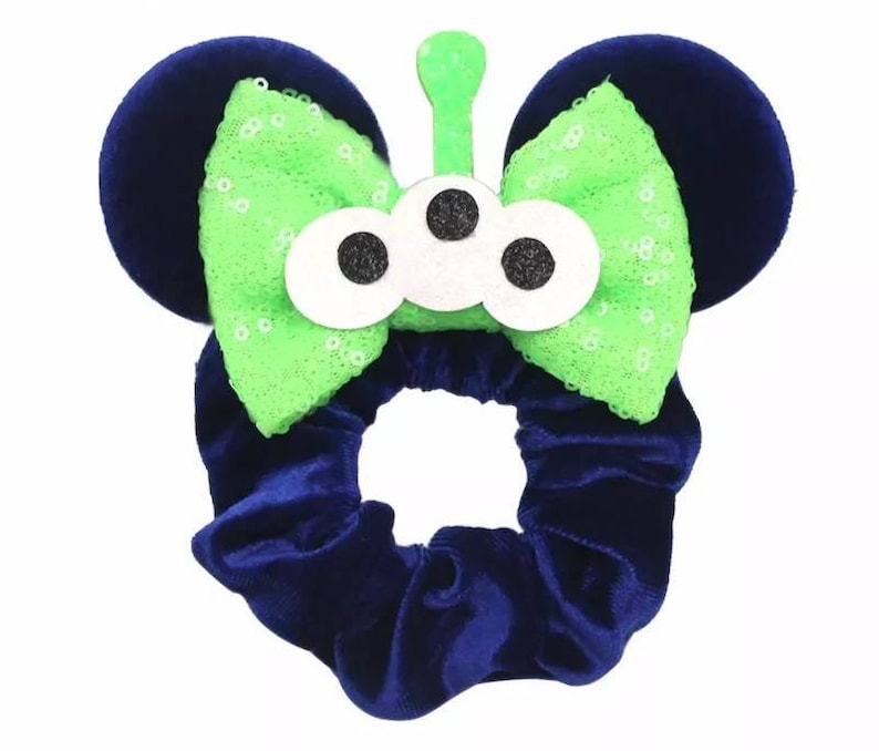 May include: A blue velvet hair scrunchie with a green and white alien-themed bow. The bow has three white eyes with black pupils and a green antenna.