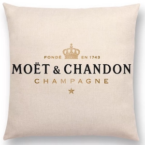 Könnte beinhalten: Ein quadratisches, cremefarbenes Dekokissen mit dem schwarz-goldenen Moët & Chandon Logo. Das Design zeigt eine goldene Krone über dem Text FONDÉ EN 1743, den Markennamen in fetten schwarzen Buchstaben und das Wort CHAMPAGNE mit einem kleinen Stern.