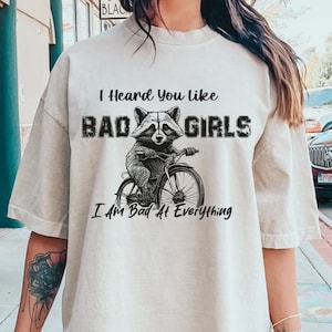 Puede incluir: Una camiseta beige claro con un gráfico negro de un mapache montando una bicicleta. El texto "I Heard You Like Bad Girls I Am Bad At Everything" está impreso en la camiseta.