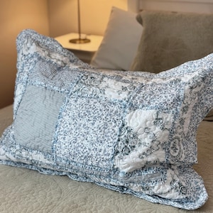 May include: A patchwork cushion with a blue and white floral and striped design. The cushion rests on a bed with a neutral-toned quilt. A lamp and other pillows are visible in the background.