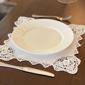 Può includere: Un piatto in ceramica bianca con disegni floreali in rilievo poggia su un centrino di pizzo bianco. Una forchetta e un coltello d'argento sono posizionati su un tavolo di legno marrone scuro, creando un'elegante mise en place. Il centrino ha un motivo delicato.