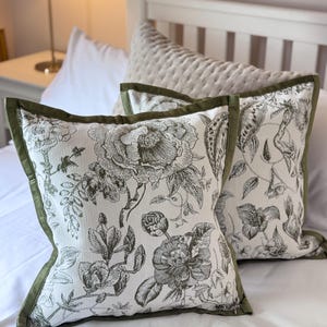 Puede incluir: Dos cojines cuadrados con una base de tela blanca y un estampado floral botánico negro detallado. Cada cojín está acabado con un borde de tela verde oliva sólido a lo largo de los lados, creando un marco distintivo para el complejo diseño floral.