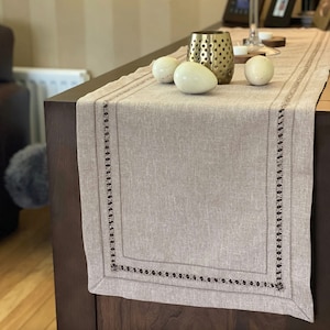 May include: A beige linen table runner with a brown embroidered border. The runner is decorated with three white eggs and a gold metal candle holder.