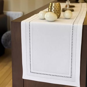 Op de afbeelding: Een witte rechthoekige tafelloper met een zwarte gestippelde rand. De loper ligt op een donkerbruin oppervlak en is versierd met witte decoratieve bollen en een goudkleurige vaas.